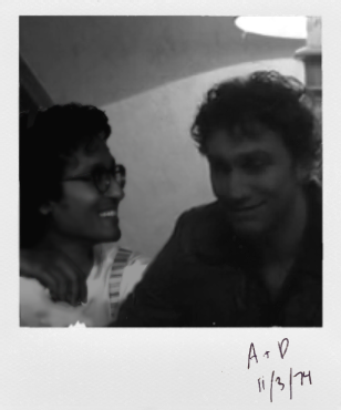 Black and white Polaroid photo showing a grinning Assad in conversation with an also-smiling Luke as Daniel Molloy in a dingy, poorly lit room. On the polaroid frame 'A + D, 11.3.74' is written in ballpoint pen.